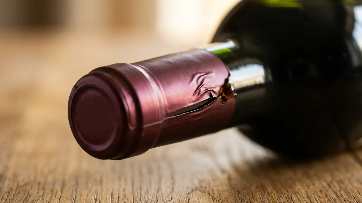 Macro close-up of a burgundy foil capsule on a green wine bottle neck with its lower edge slightly lifted and creased