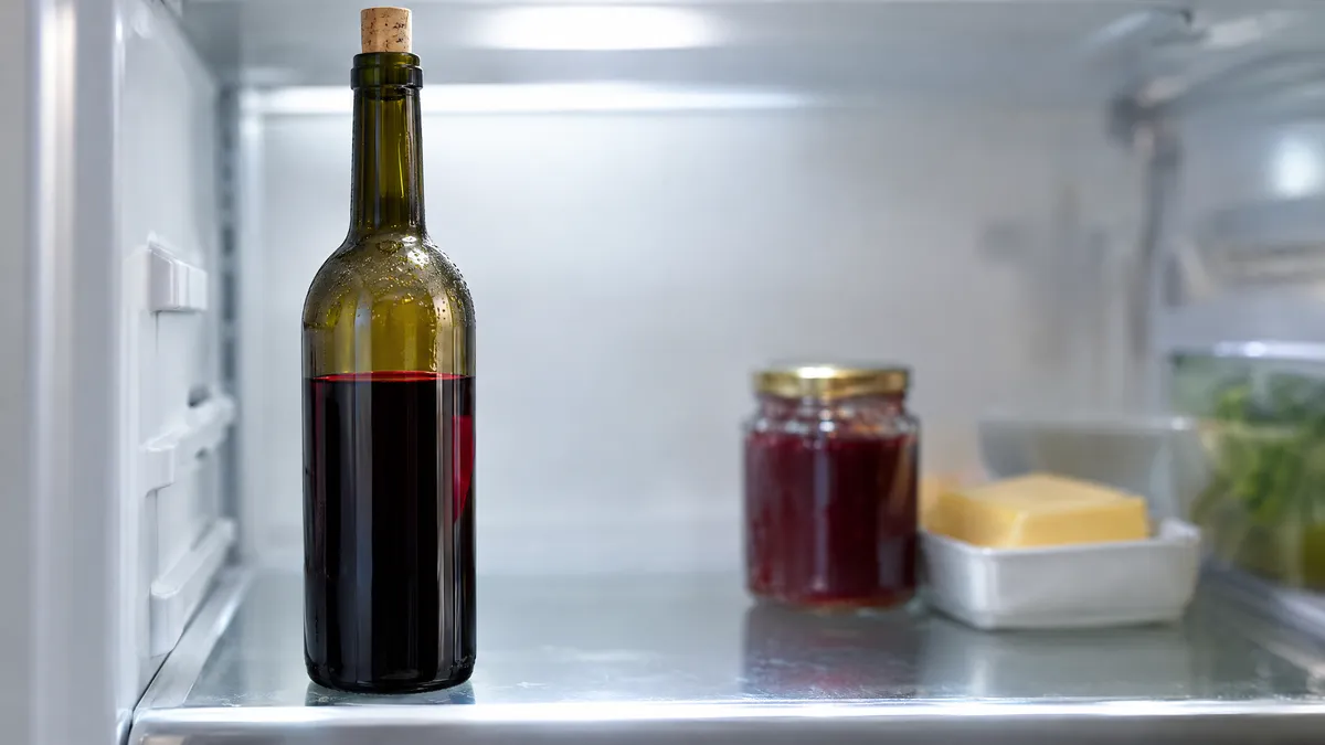 A half-full bottle of red wine with its cork pushed back in, standing upright on a refrigerator shelf