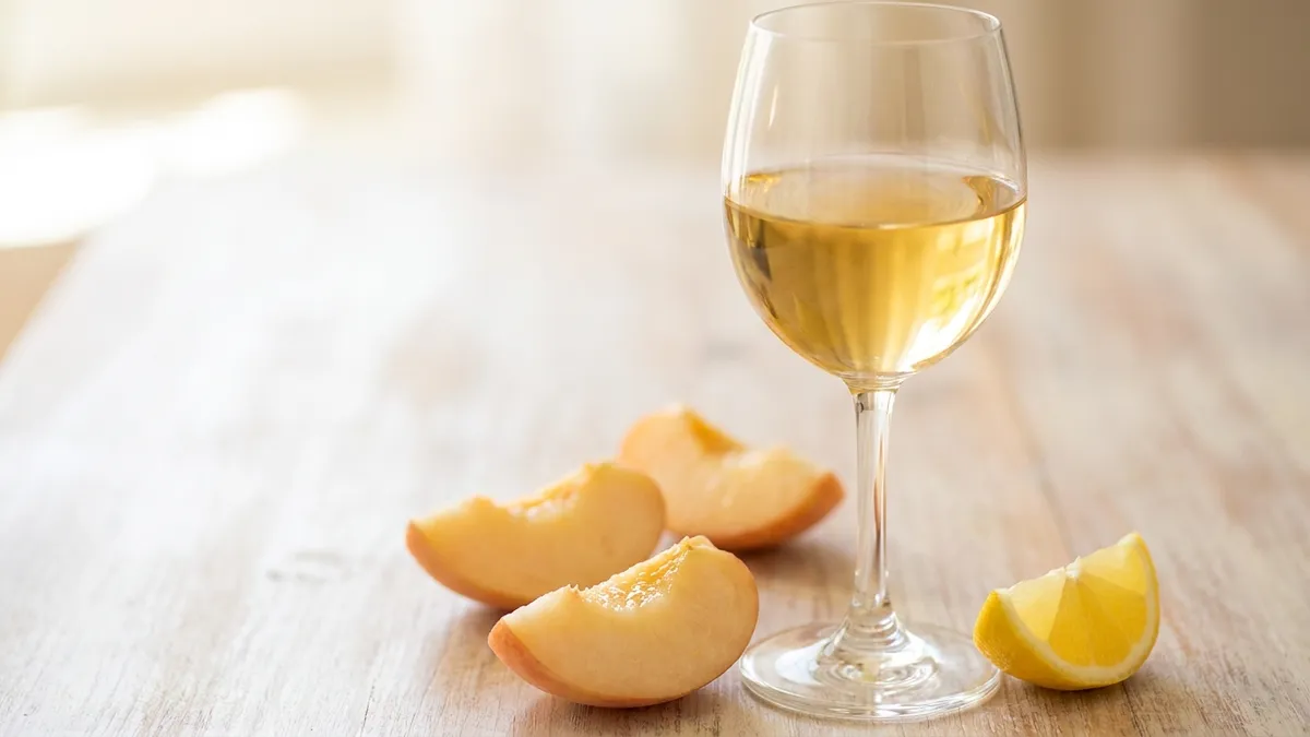 A glass of golden white wine beside sliced stone fruit and a lemon wedge on a light wooden surface