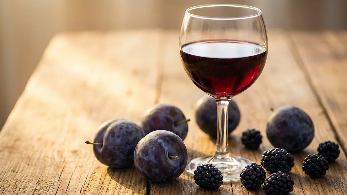 A glass of deep-colored red wine on a wooden table beside scattered blackberries and plums