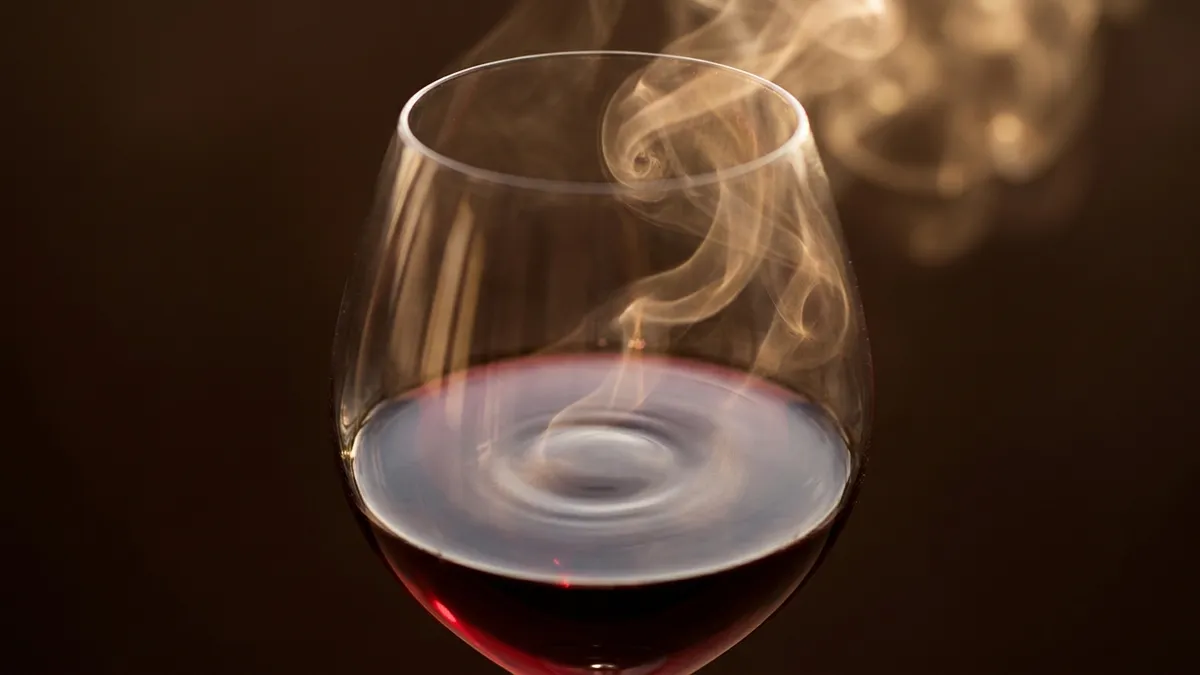 A top-down view into the bowl of a wine glass holding deep ruby-garnet wine, with a soft luminous vapor of aromatic compounds rising in delicate ribbons from the wine surface