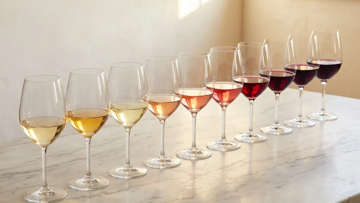 Ten wine glasses arranged on a marble surface ranging from pale straw-gold whites to deep ruby-purple reds, lit by warm overhead daylight