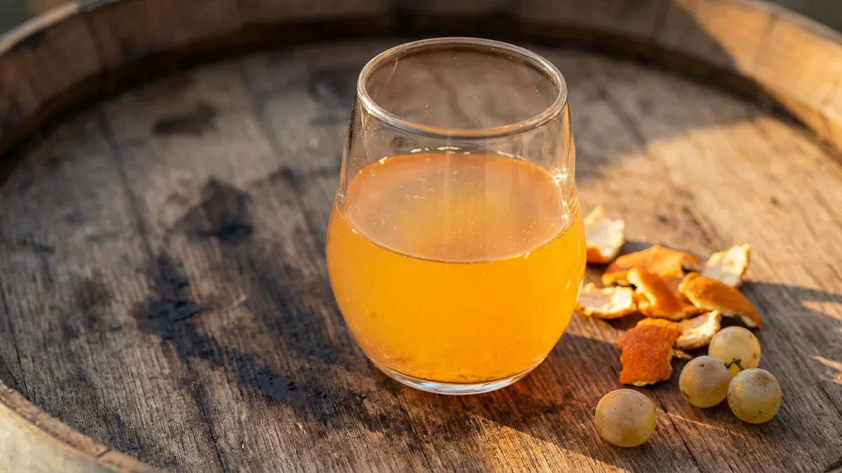 Glass of amber-colored orange wine next to white grapes on a rustic wooden surface