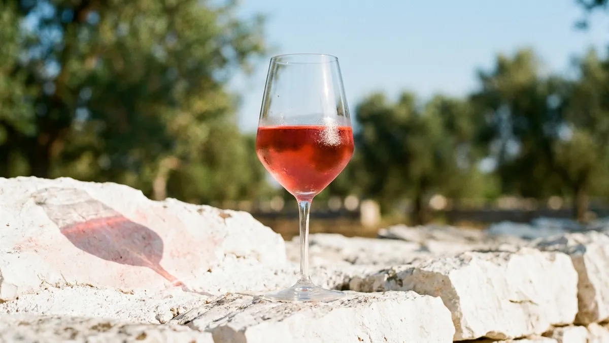 A glass of deep coral rosato standing on a white limestone wall with olive trees and a pale sky behind