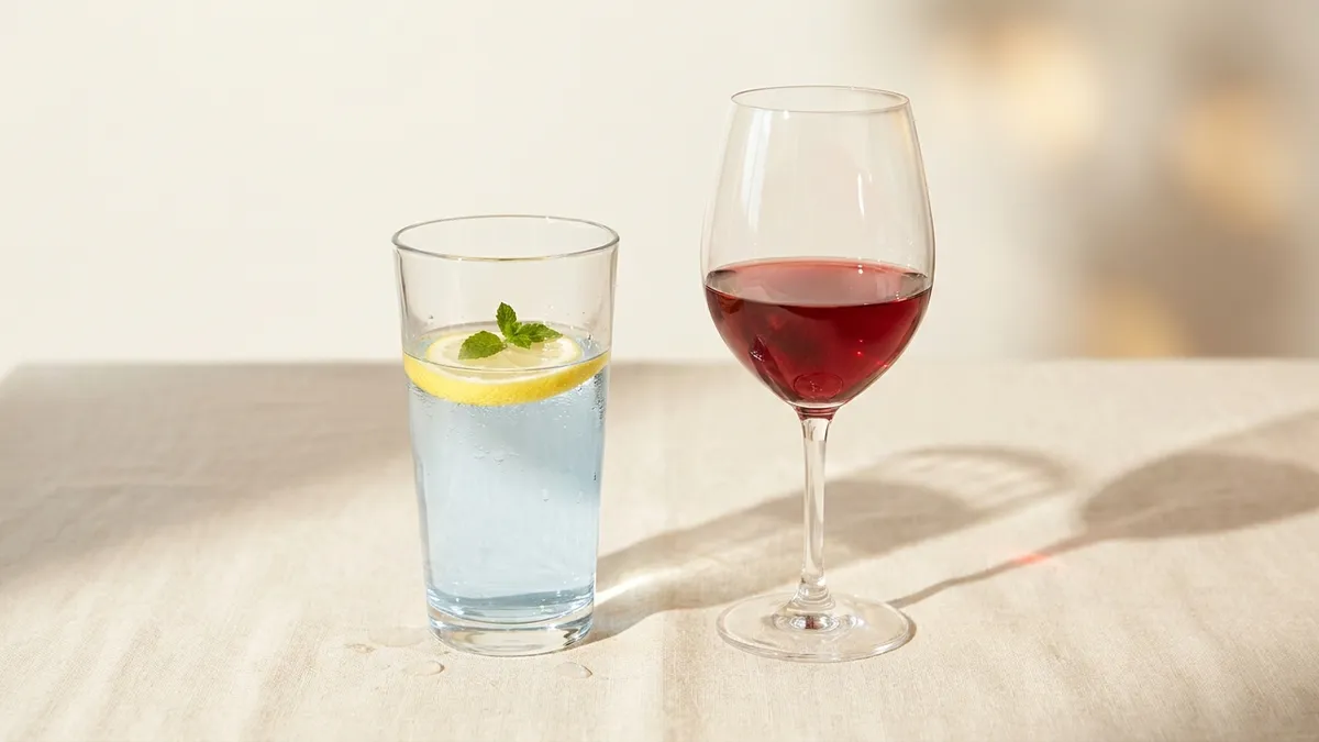 A glass of wine and a glass of sparkling water on a dinner table, side by side, with bread and olive oil between them, illustrating the alternate-water habit that paces an evening