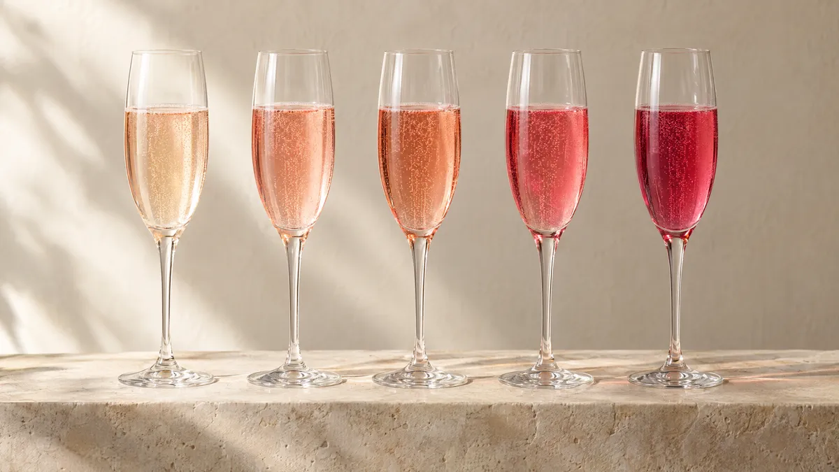 Five glasses of sparkling rosé in a row on a stone ledge, colours moving from pale onion-skin salmon to deep raspberry pink