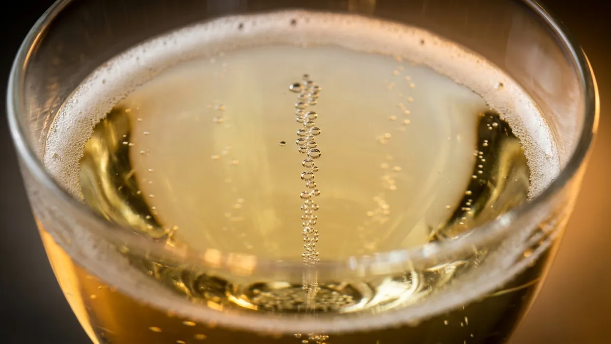 Extreme macro close-up of fine bubbles rising in a straight chain through pale gold sparkling wine in a crystal flute, backlit by warm amber light