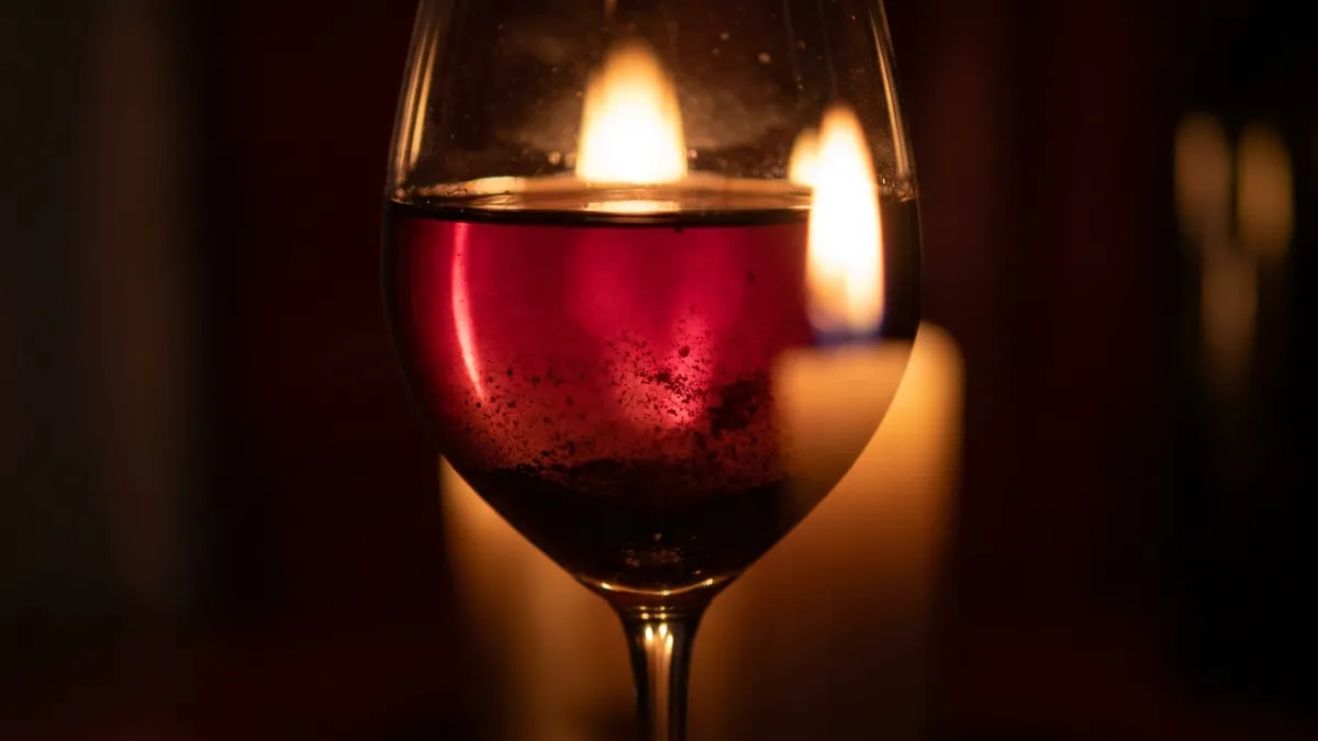 A glass of deep garnet-red aged wine held up to a lit candle, a fine dark cloud of sediment visible drifting near the bottom of the glass against the warm flame