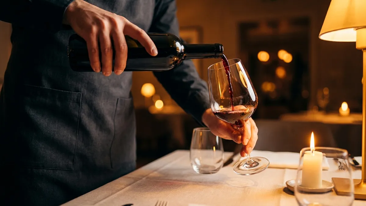 A sommelier in a dark apron pouring red wine into a glass at a restaurant table, hands and bottle in sharp focus, warm candlelight behind
