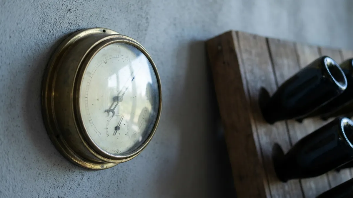 A wall-mounted analog hygrometer and thermometer hanging beside a small wooden wine rack in dim ambient light