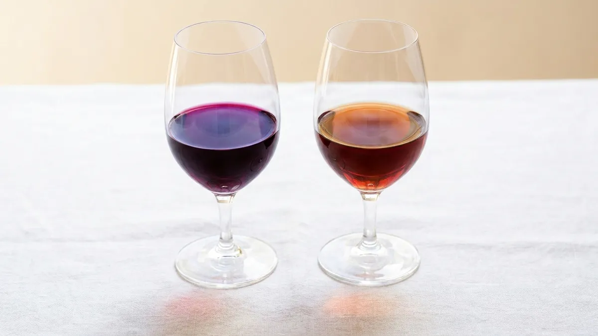 Two red wine glasses on white linen, a young ruby-purple red beside an older red that has turned brick-orange