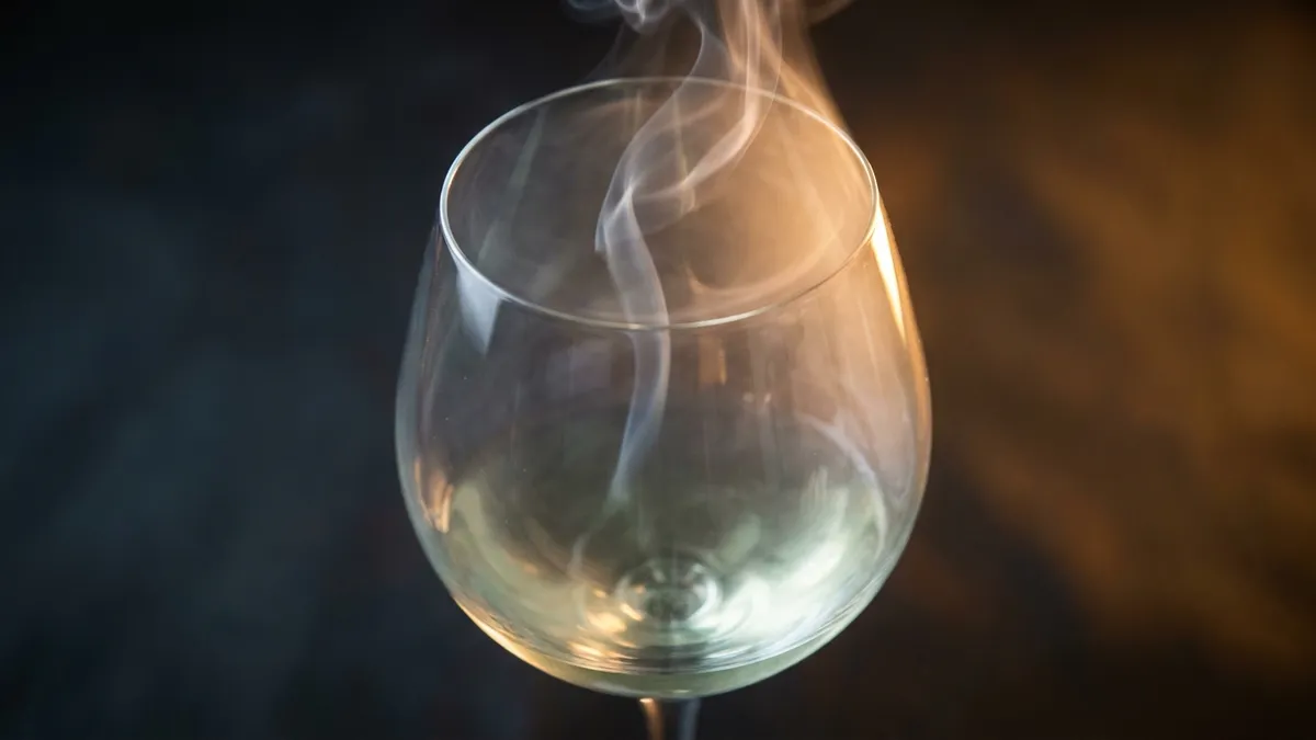 Close view into a wine glass of very pale white wine with a thin wisp of smoke rising from the bowl against a dark background