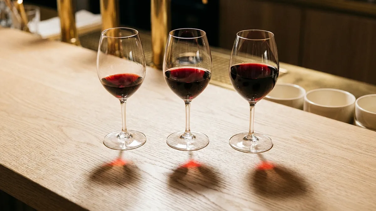 Three stemmed glasses in a row on a wooden bar, each poured to a visibly different level, from a small tasting pour to a generous fill