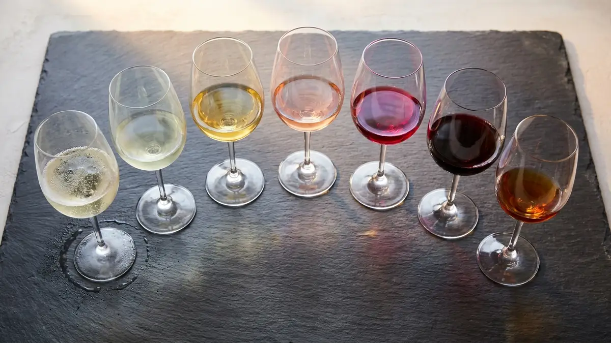 Seven wine glasses arranged in an ascending arc on dark slate, from pale sparkling on the left through golden whites, rosé, and reds to a small pour of amber dessert wine on the right