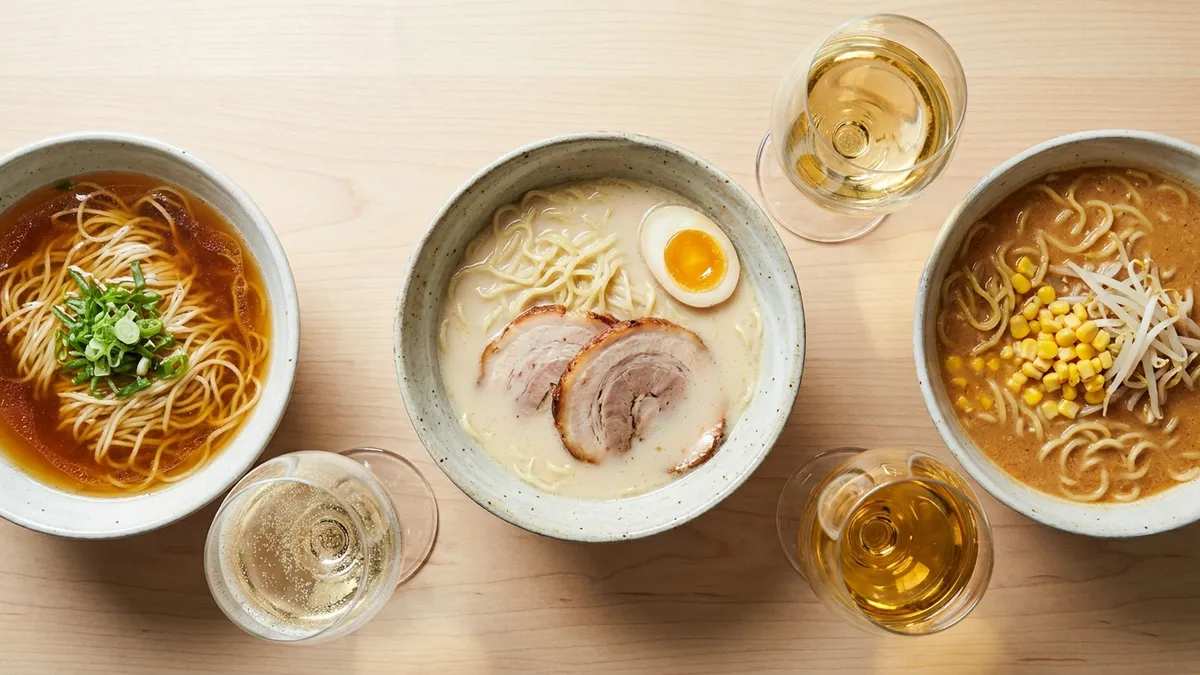 Three bowls of ramen from above, a clear amber soy broth, a creamy white pork broth and a thick tan miso broth with corn, each beside a glass of white or sparkling wine