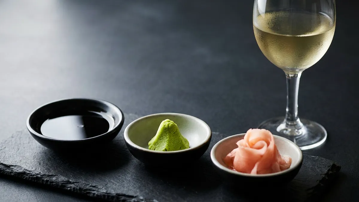 A small dish of soy sauce, a mound of bright green wasabi, and pale pink pickled ginger arranged beside a glass of crisp white wine on a dark surface