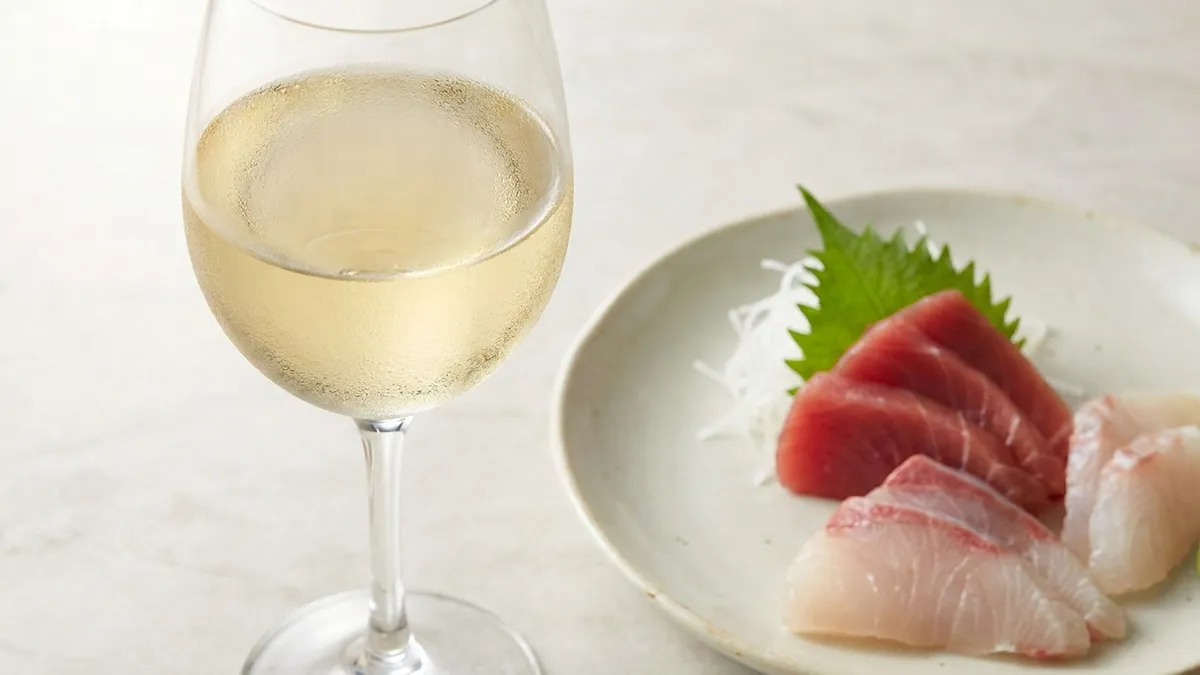 A glass of pale, chilled white wine beside an arrangement of clean, ruby-red and pale sashimi slices on a light ceramic plate in bright soft light