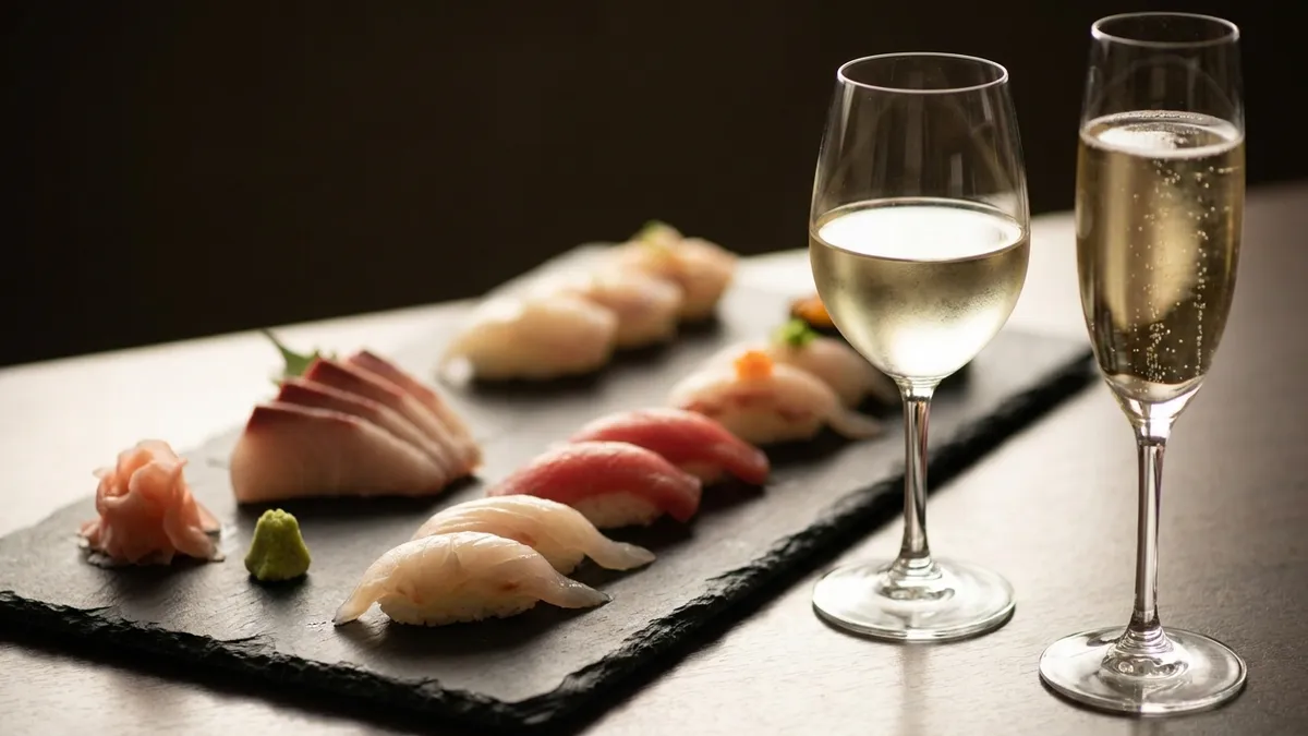 An elegant sushi spread of nigiri and sashimi on a dark slate board beside two glasses of pale, chilled white and sparkling wine in soft warm light