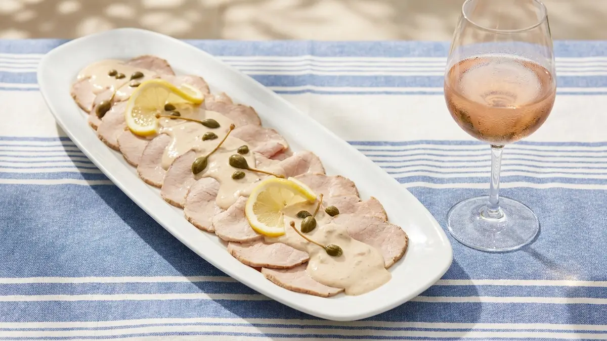 Thin slices of cold veal under a creamy tuna sauce with capers on a platter beside a glass of pale pink rosé